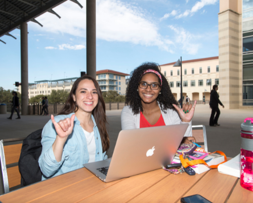 Two students outside in front of a laptop doing the birds up hand sign