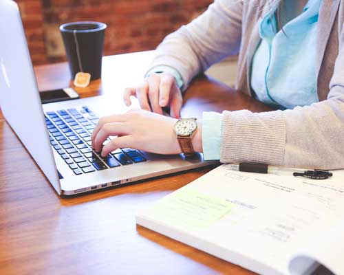 Person typing on a laptop at a wooden table with a coffee cup and documents