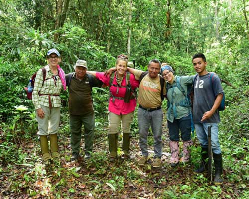 Group of six people posing in a lush forest, wearing outdoor clothing and boots.