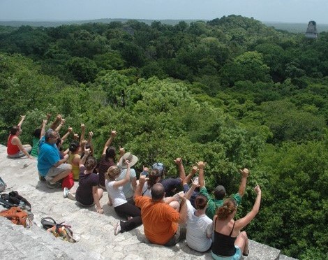 A group of people sitting on a stone structure, overlooking a dense forest with a distant ancient ruin visible